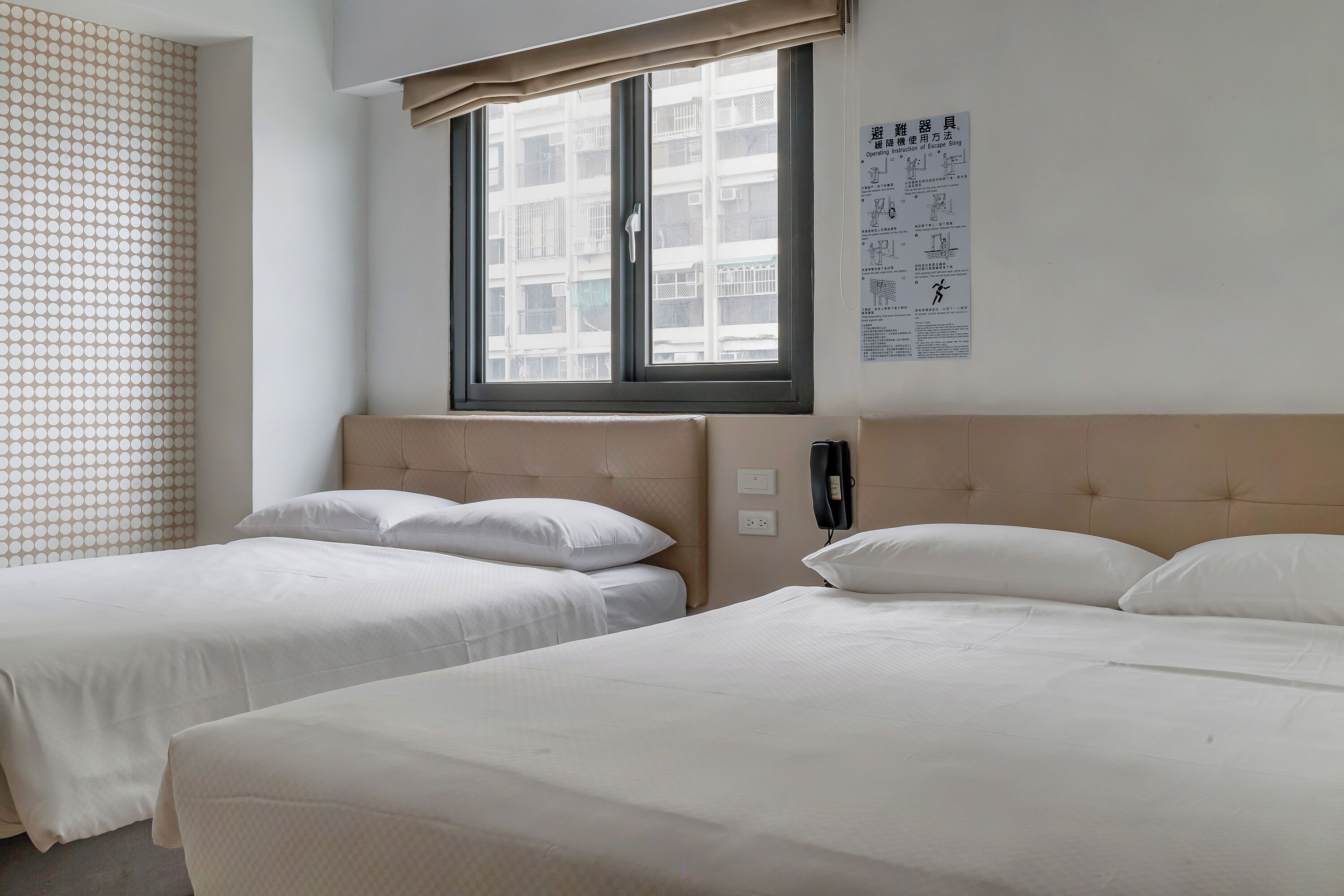 A hotel room with two single beds, a large window with blinds, a wall-mounted phone, and a patterned curtain.