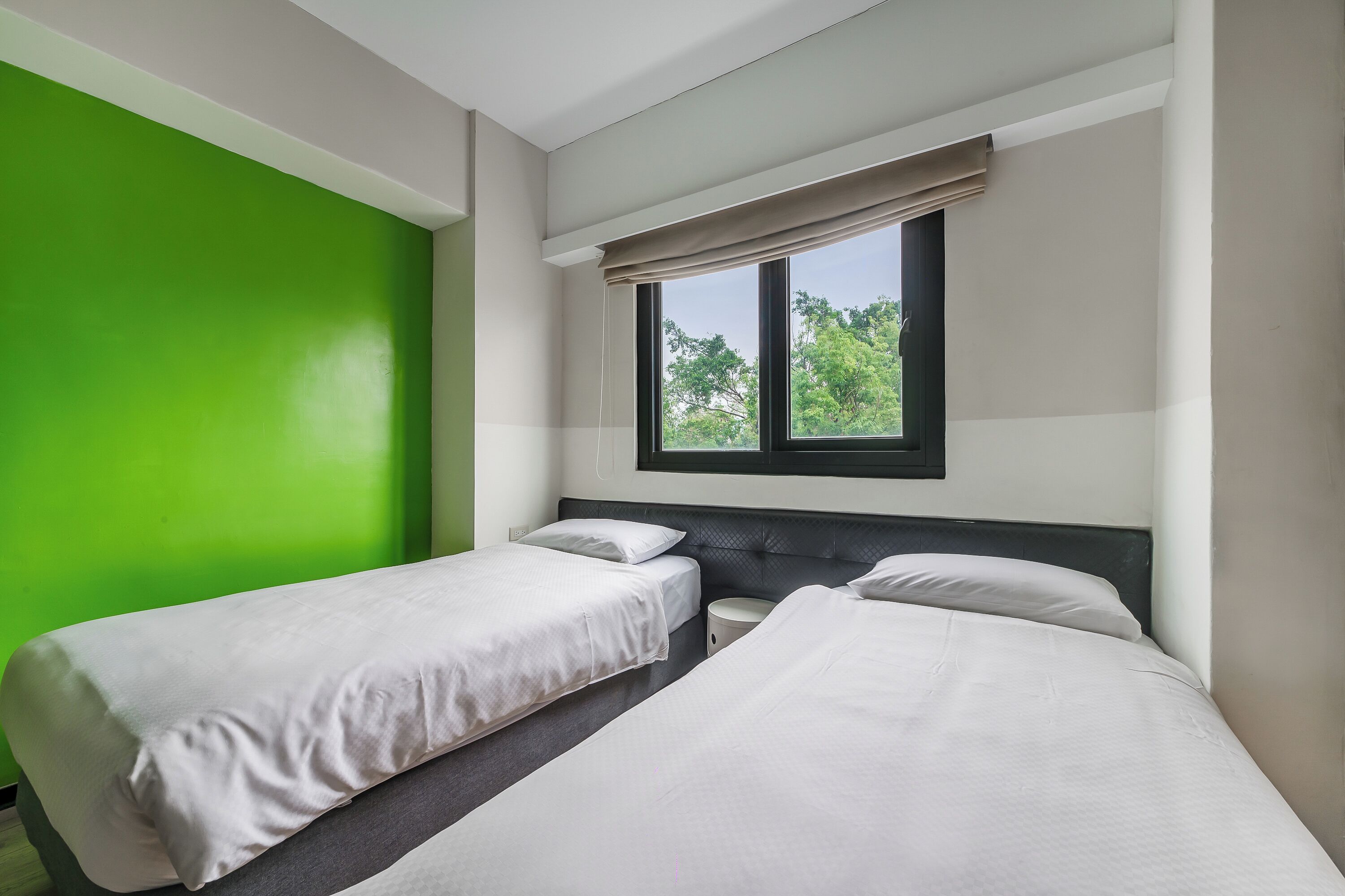 A room with two beds, a green wall, and a window with a view of trees.
