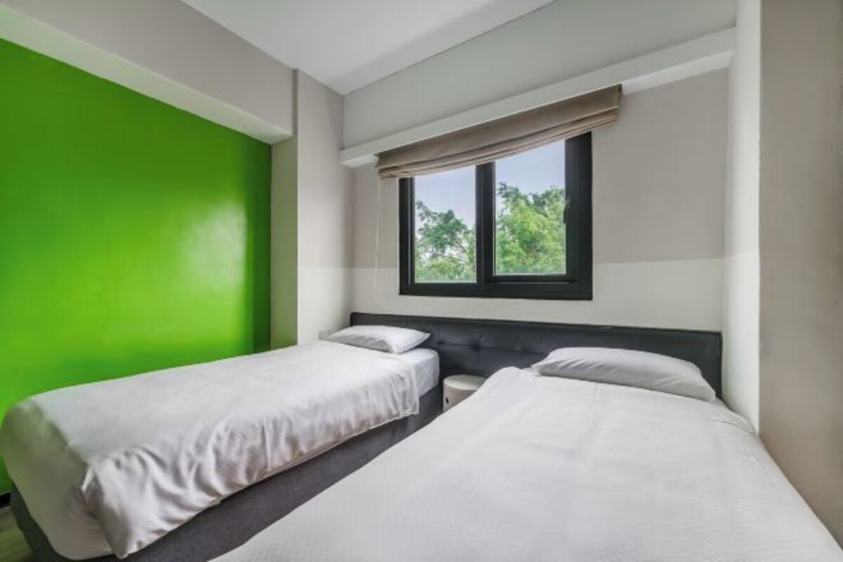 A hotel room with two single beds, a window with a view of greenery, and a green accent wall.