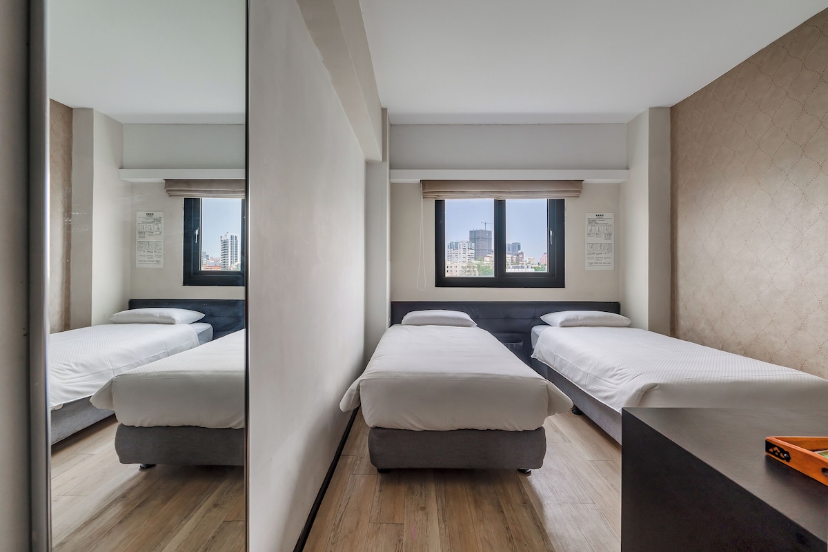 A hotel room with two single beds, a wooden floor, a large window with a city view, and a mirror reflecting the room.
