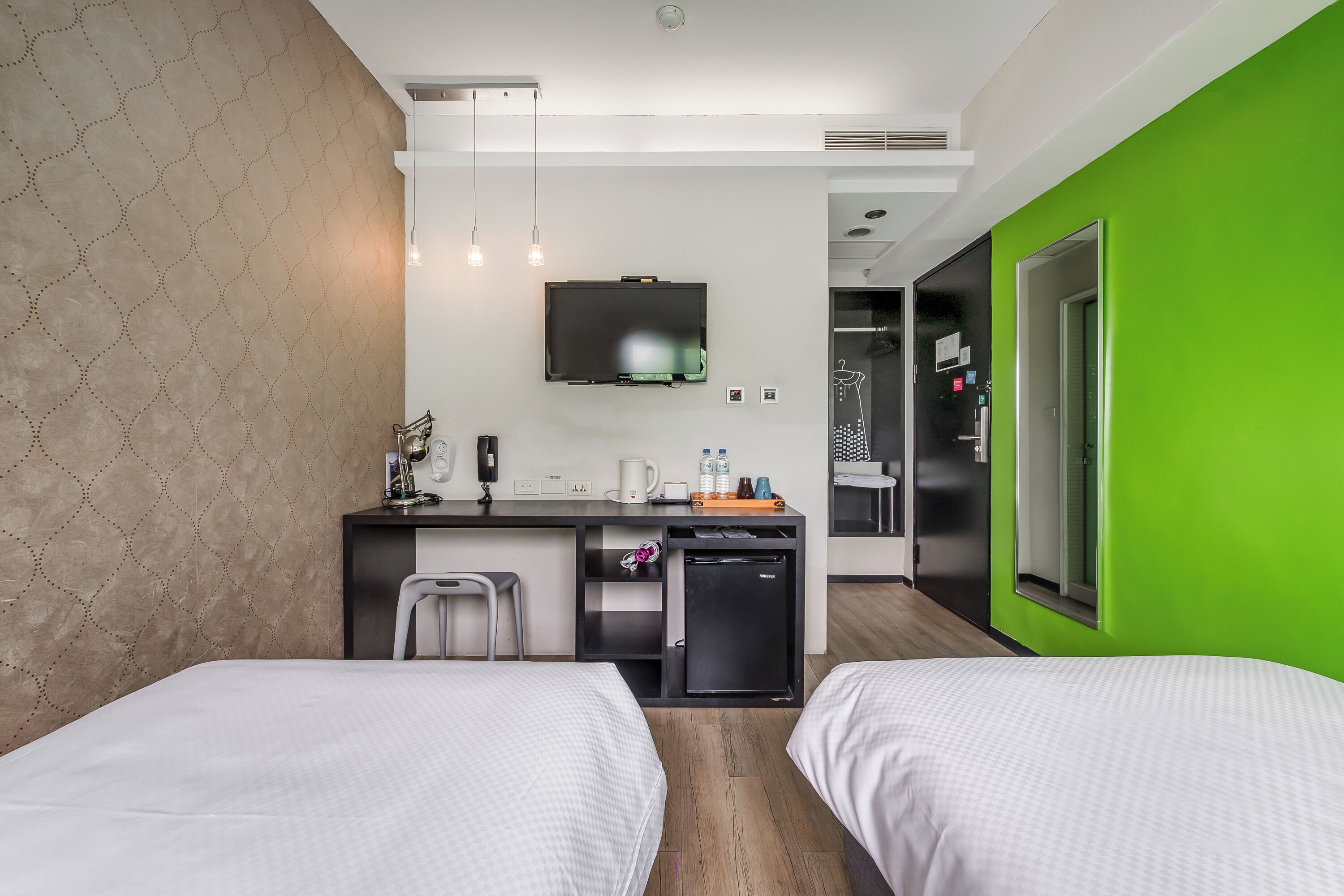 A modern hotel room with two beds, a kitchenette, a TV, and a green accent wall.