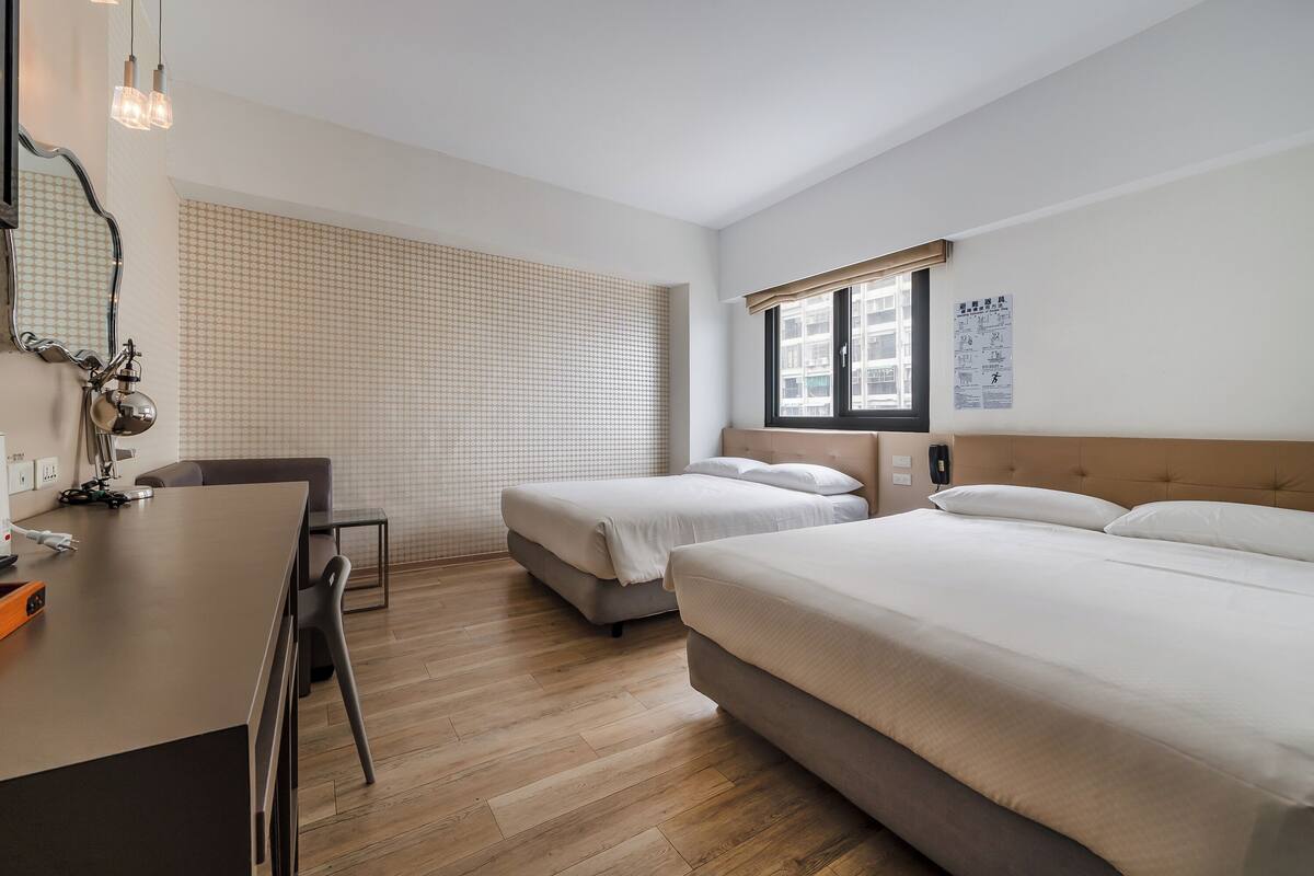 Une chambre d’hôtel avec deux lits, un bureau et une chaise. Il y a une fenêtre donnant sur les bâtiments environnants.