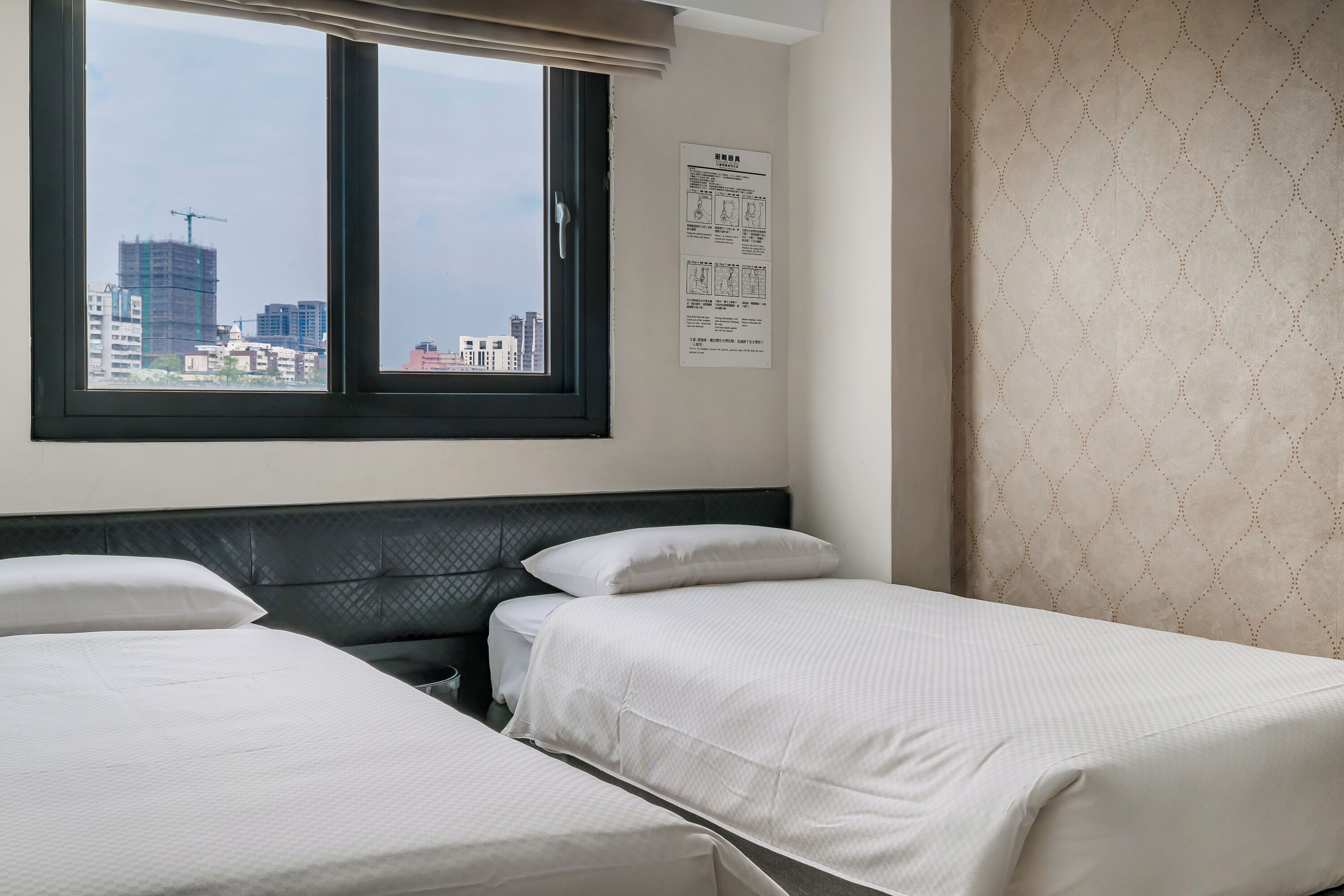 Quarto de hotel com duas camas, uma janela com vista para prédios e um mural na parede.