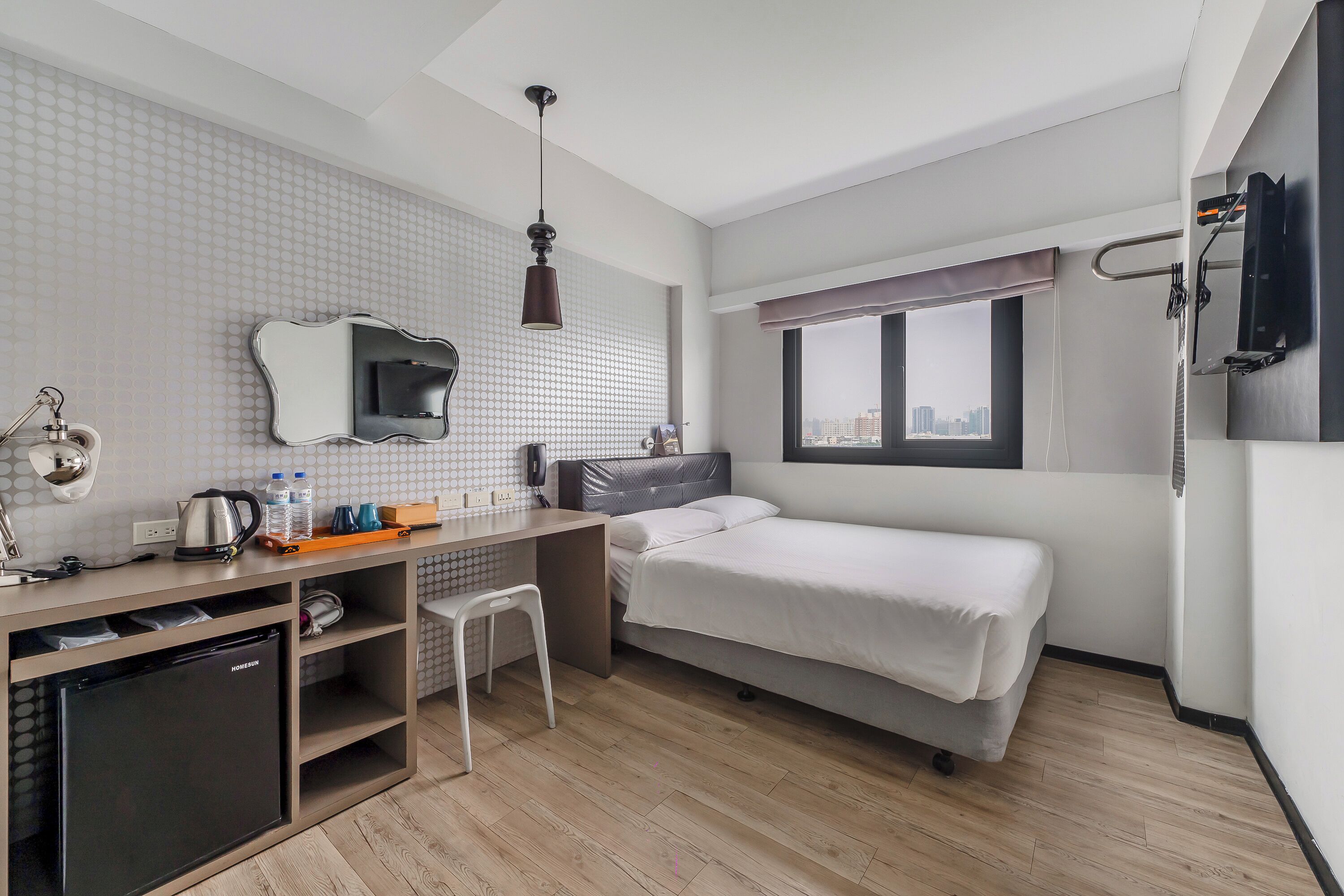 Habitación de hotel compacta con cama, escritorio con silla, refrigerador pequeño, espejo y ventana con vista a la ciudad.