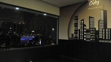 A hotel room with a large window offering a city view at night, a wall mural of a city skyline, and a sign that reads "City Travel in Lia's inn".