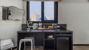 Habitación de hotel con escritorio, una silla, un refrigerador pequeño, un espejo y ventana con vista a la ciudad.