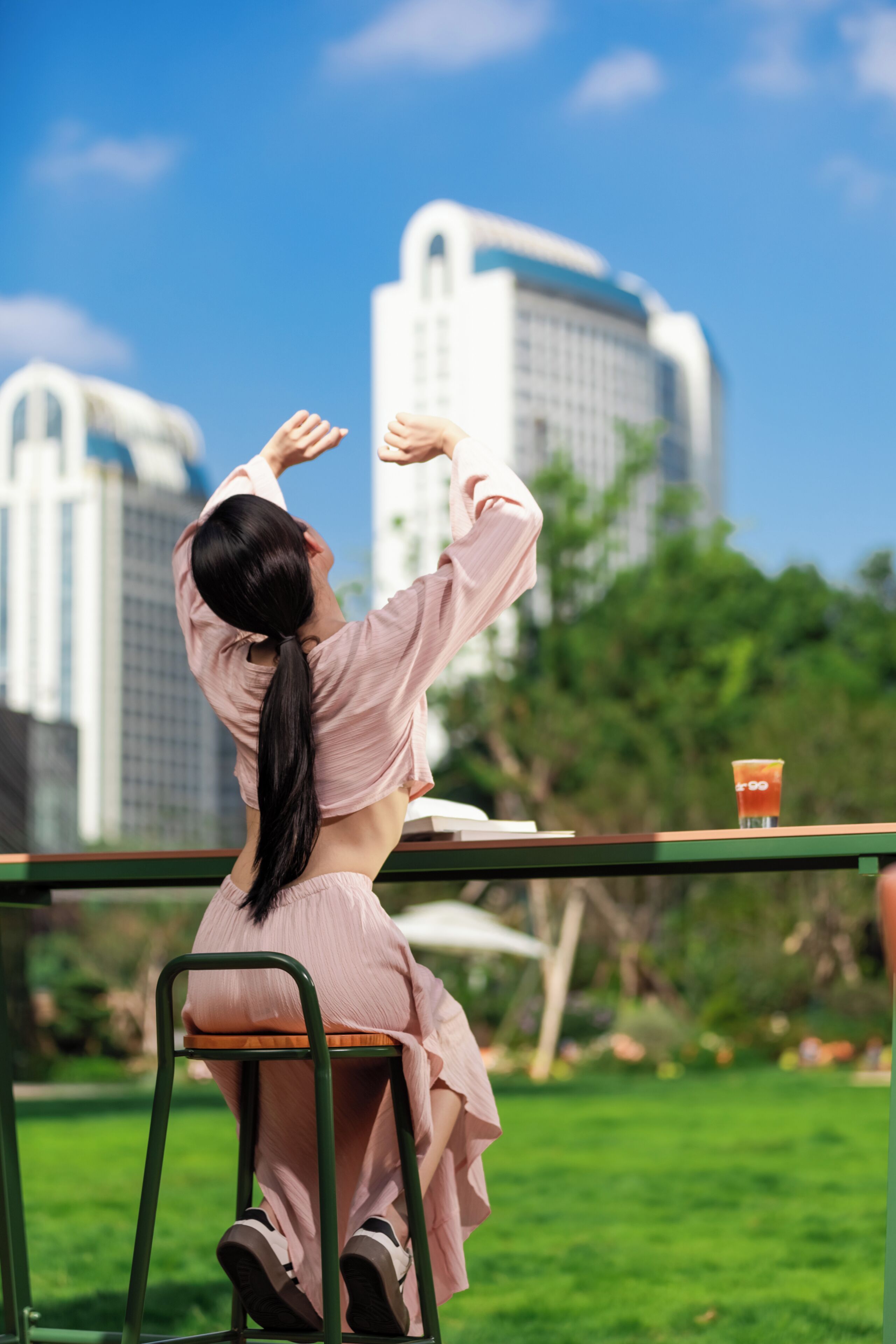 A person sitting on a chair outdoors with a table in front, a drink on the table, and buildings in the background.