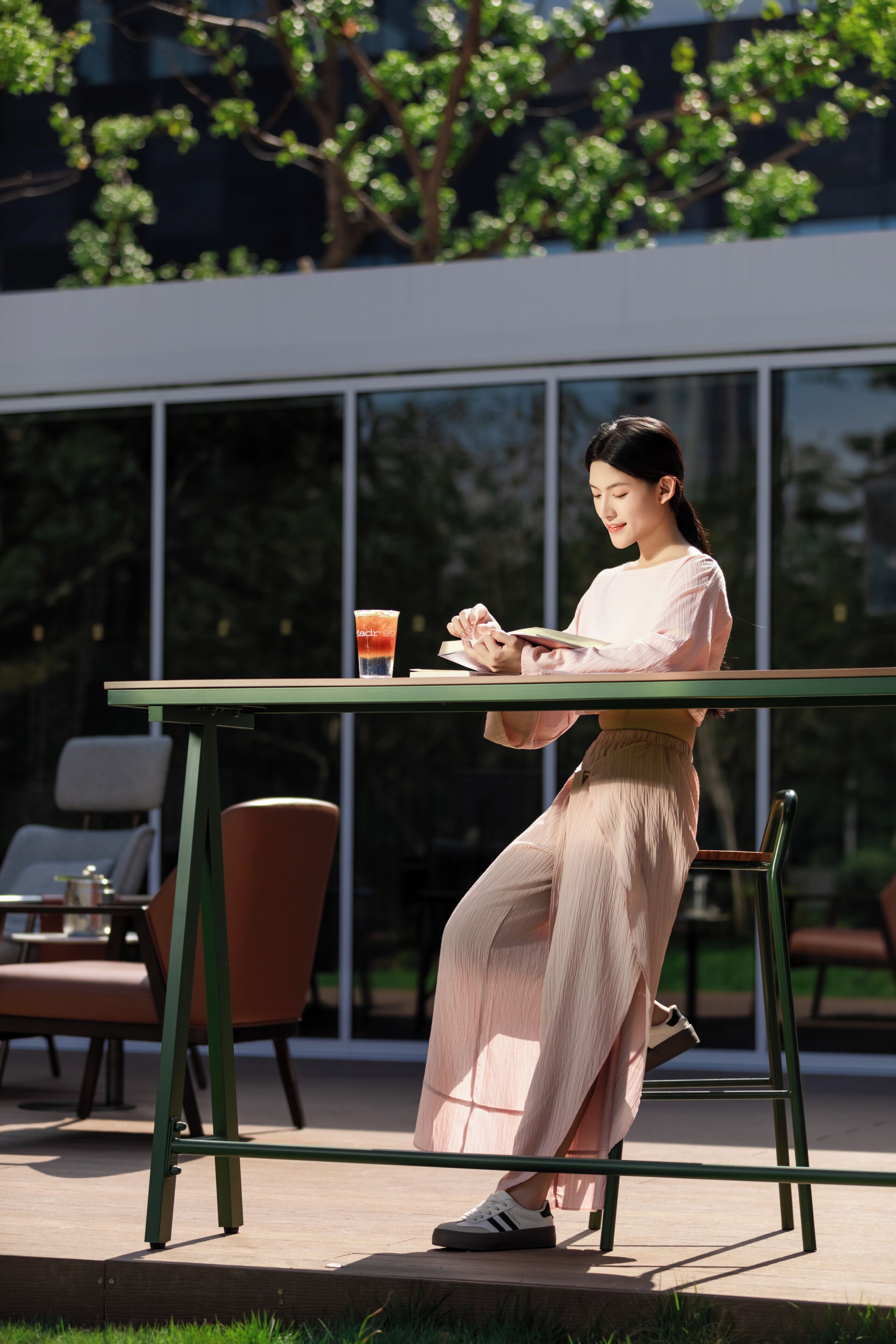 Una persona sentada en una mesa al aire libre, con una bebida, frente a un edificio moderno con grandes ventanales.
