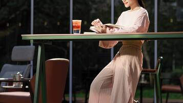 Una persona sentada en una mesa al aire libre, con una bebida, frente a un edificio moderno con grandes ventanales.