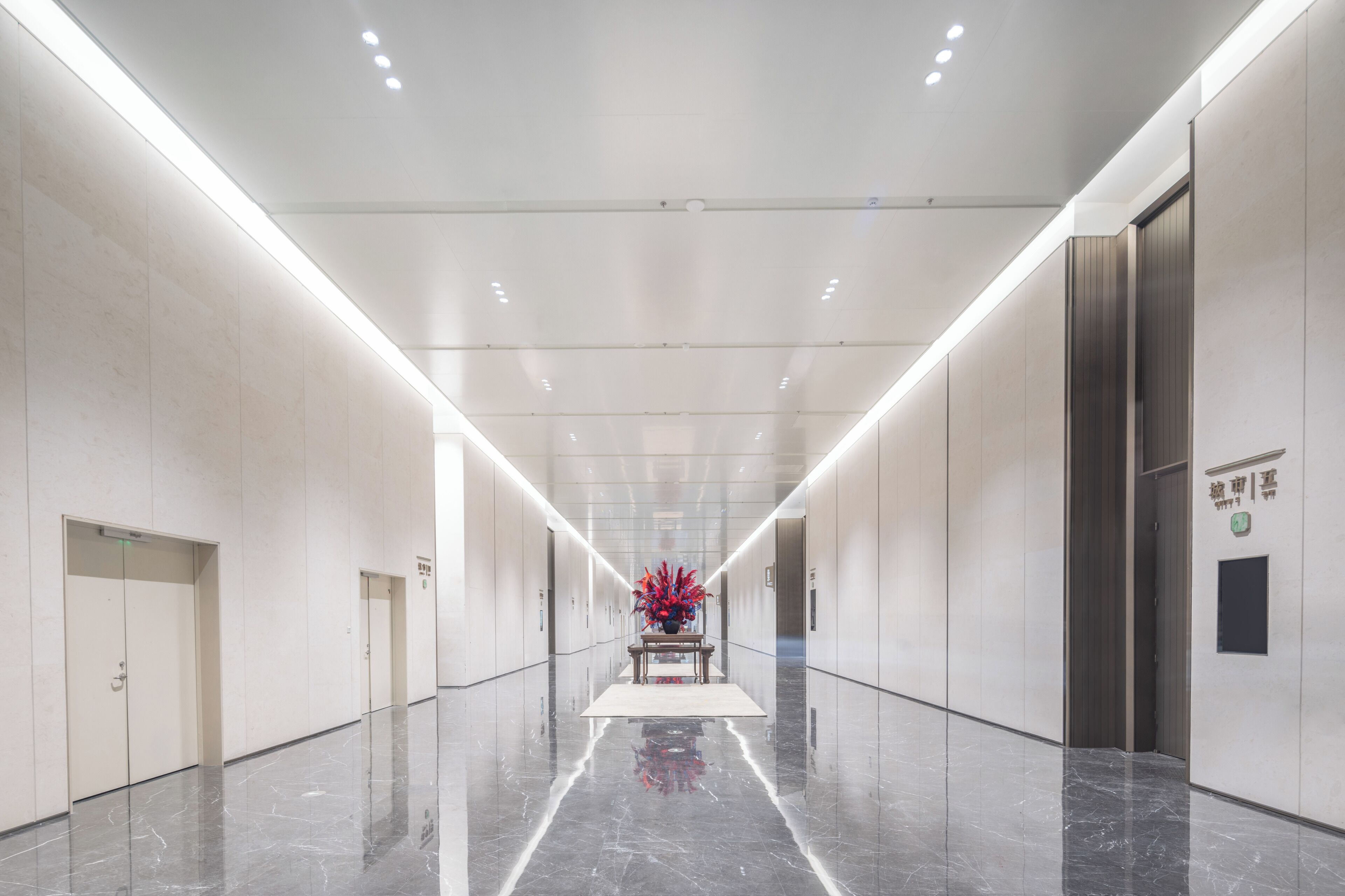 A long, well-lit hallway with a reflective floor, doors on both sides, and a central table with a red floral arrangement.
