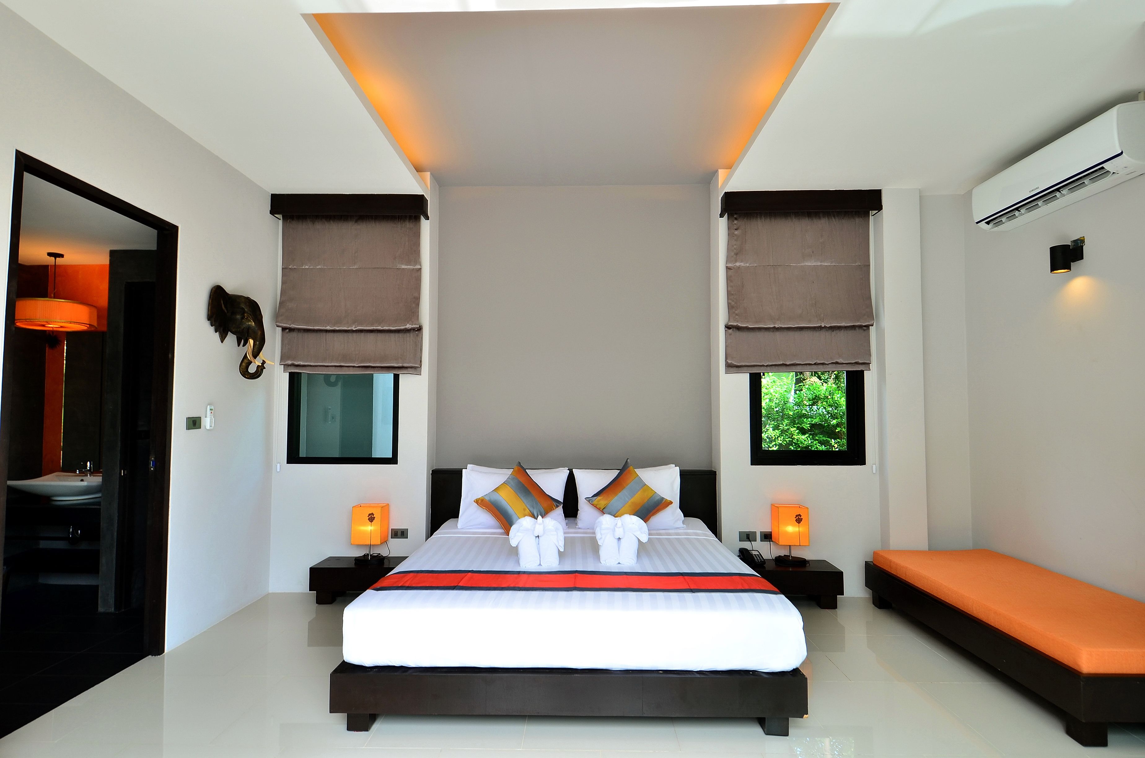 Una habitación de hotel moderna con una cama grande, dos mesitas de noche, un banco y vistas al exterior con vegetación.