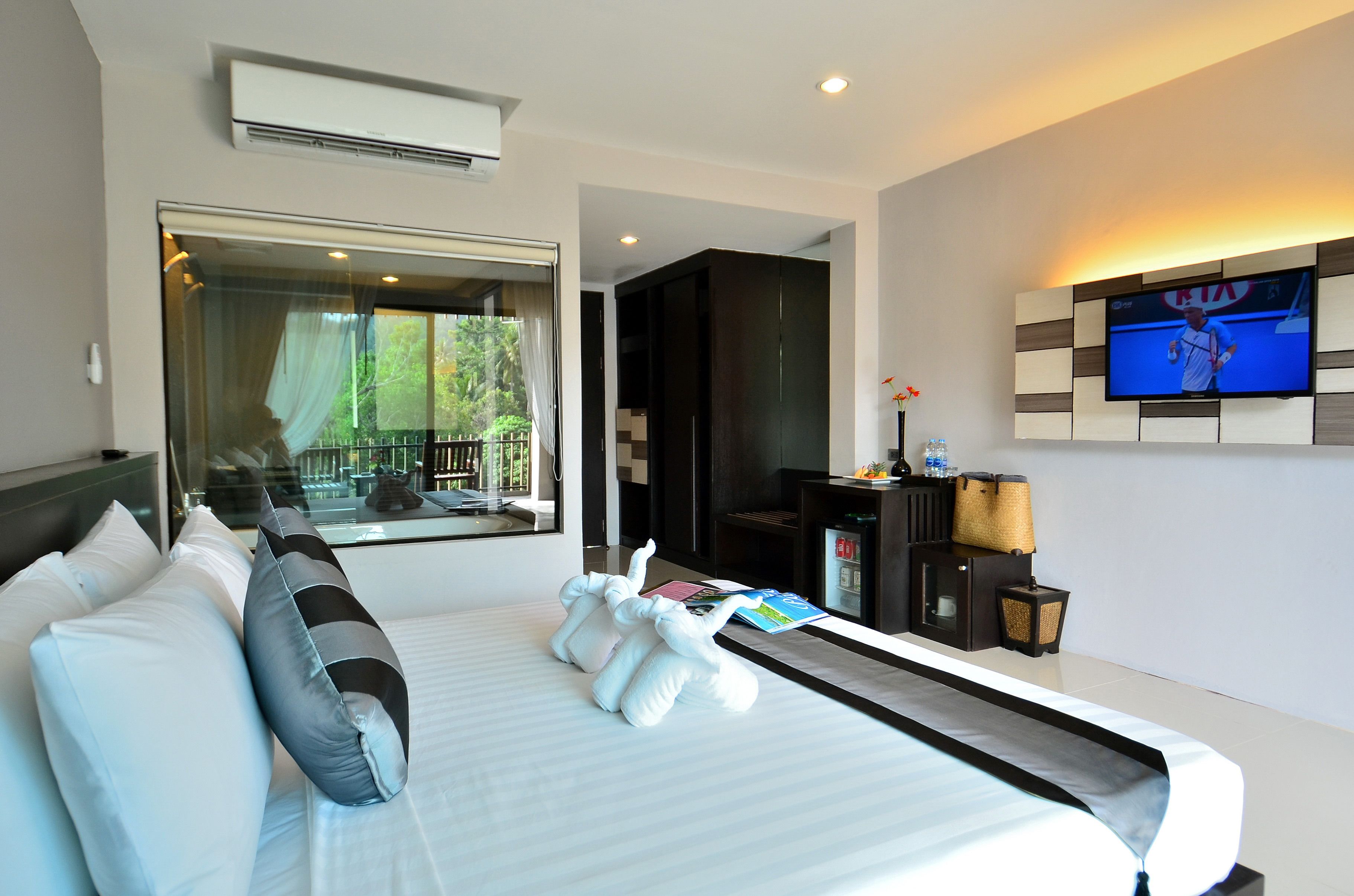 A hotel room with a large bed, a TV mounted on the wall, a window with a view of greenery, and a minibar.
