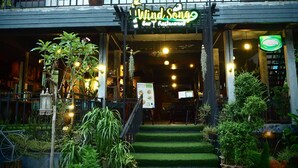 Un restaurante llamado 'Wind Song Bar & Restaurant' con un área de asientos al aire libre cubierta, plantas en macetas y una escalera que conduce a la entrada.