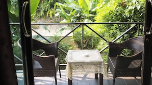 Un balcón con sillas de mimbre y una mesa, con vistas a un jardín exuberante y una casa tradicional.