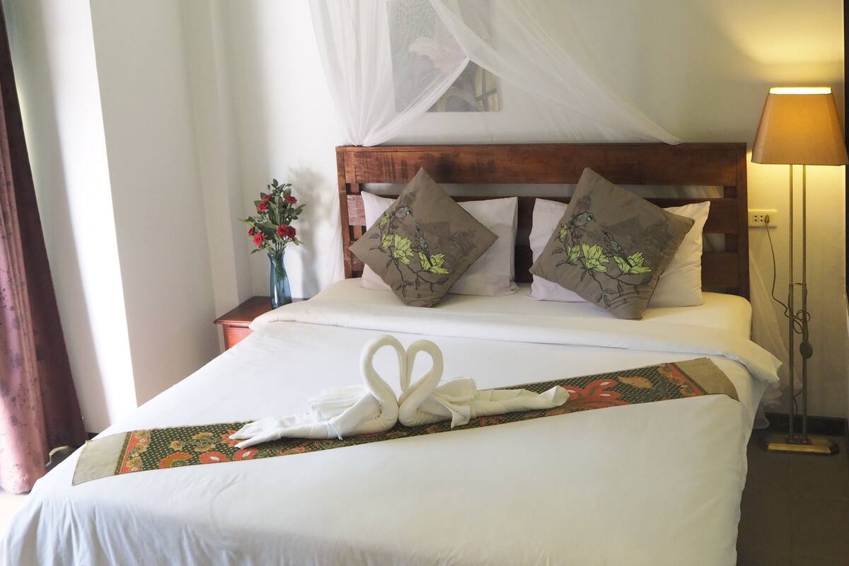 Una cama bien hecha con una colcha blanca, dos almohadas decorativas, una mesita de noche con un florero y un ventilador de pie.