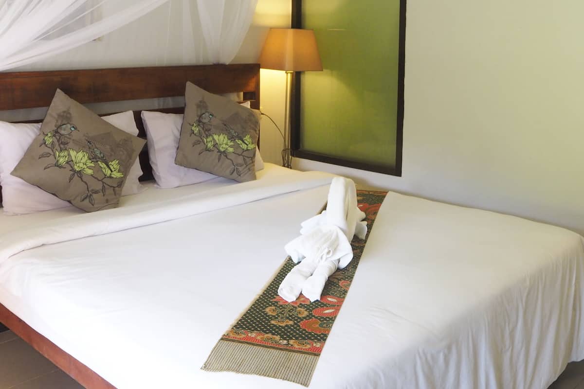 Una cama bien hecha con edredón blanco, dos almohadas decorativas, mosquitero, lámpara de noche y aire acondicionado de pared.