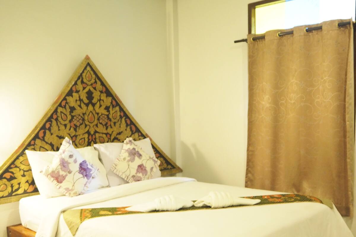 Una cama bien hecha con ropa de cama blanca y almohadas florales, un cabecero de madera con un diseño decorativo y una cortina marrón en una ventana.