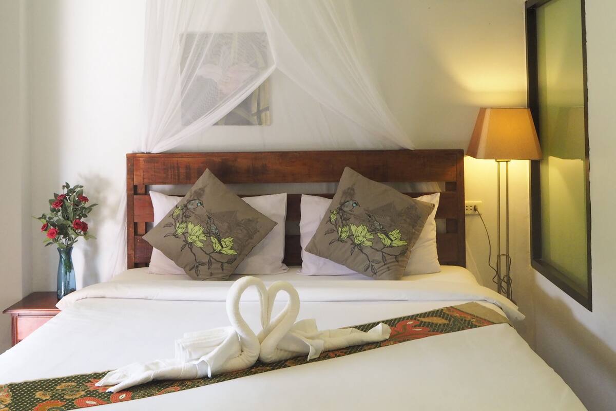 Habitación de hotel con una cama, una mesita de noche con un florero y un aire acondicionado de pared, además de una lámpara de noche.