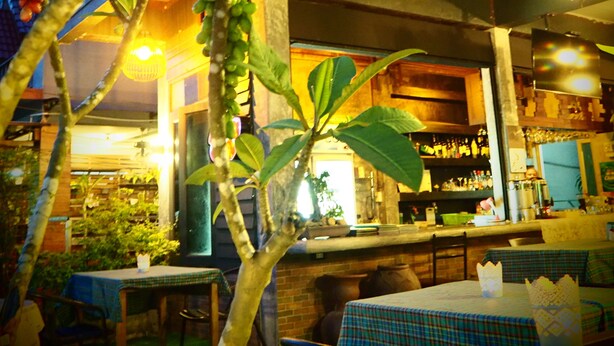 Un área de comedor al aire libre con mesas y sillas, rodeada de follaje y una zona de bar con estantes llenos de botellas.