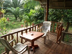 A wooden deck with a table and chairs, surrounded by lush greenery and a thatched roof structure.