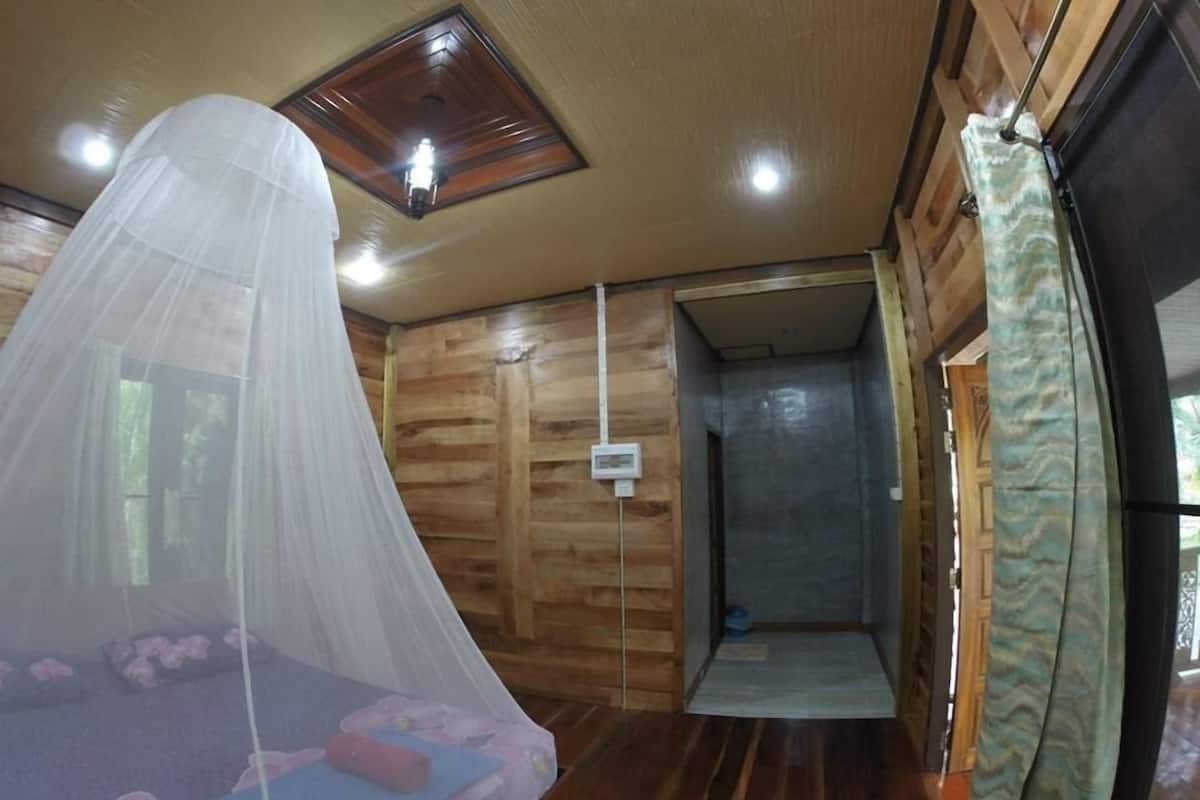 A wooden cabin room with a bed under a mosquito net, a shower area, and a wooden ceiling.