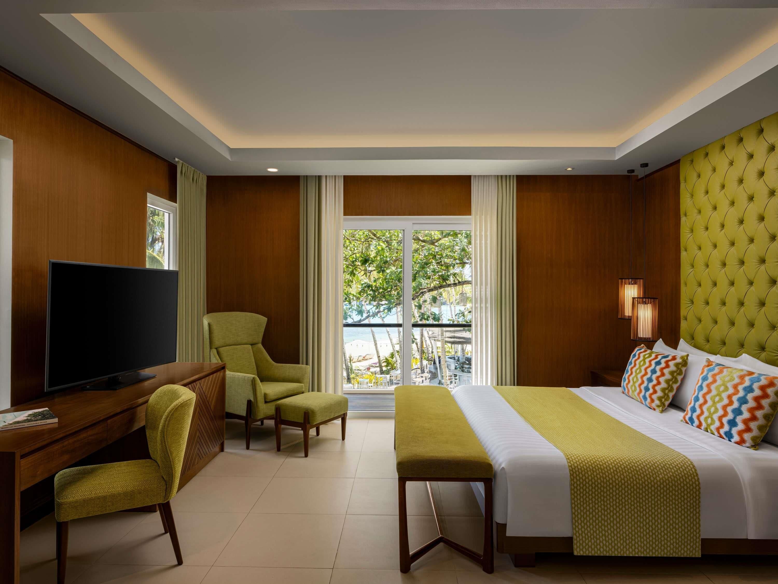 A hotel room with a bed, a desk with a TV, a chair, a bench, and a view of the outdoors.
