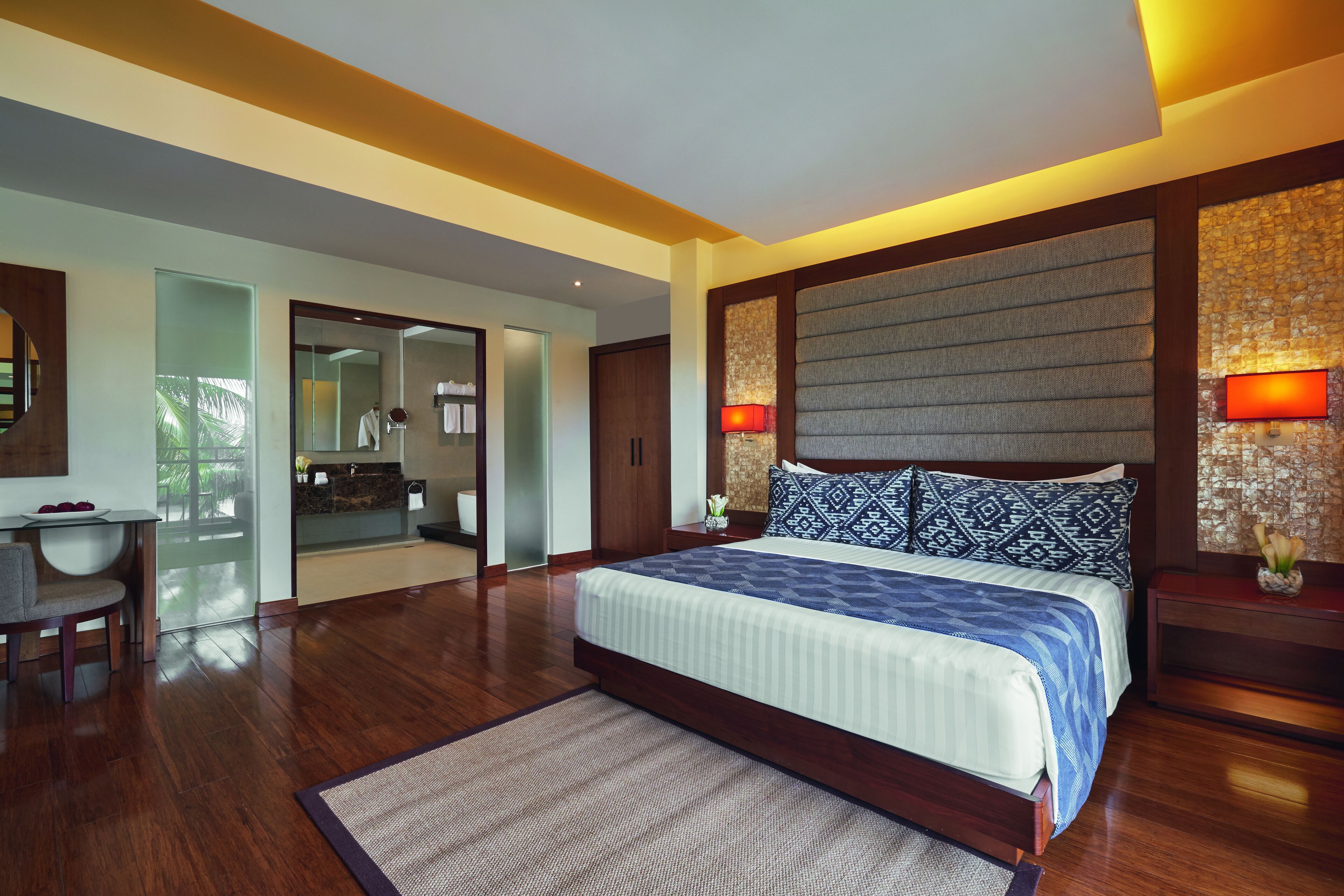 A modern bedroom with a large bed, wooden flooring, a desk, and a bathroom visible through a glass door.
