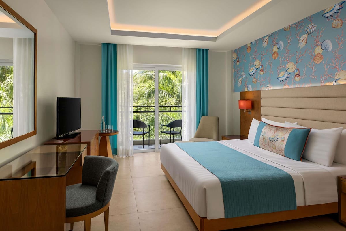 Habitación de hotel con cama, escritorio con televisor, una silla, balcón con sillas y vistas a zonas verdes.