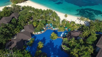 An aerial view of a resort with a central swimming pool, surrounded by palm trees and a beach.
