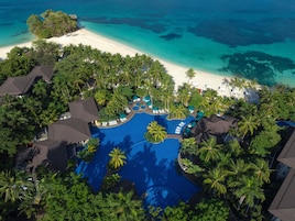 Vista aérea de un resort con una piscina central, rodeado de palmeras y una playa.