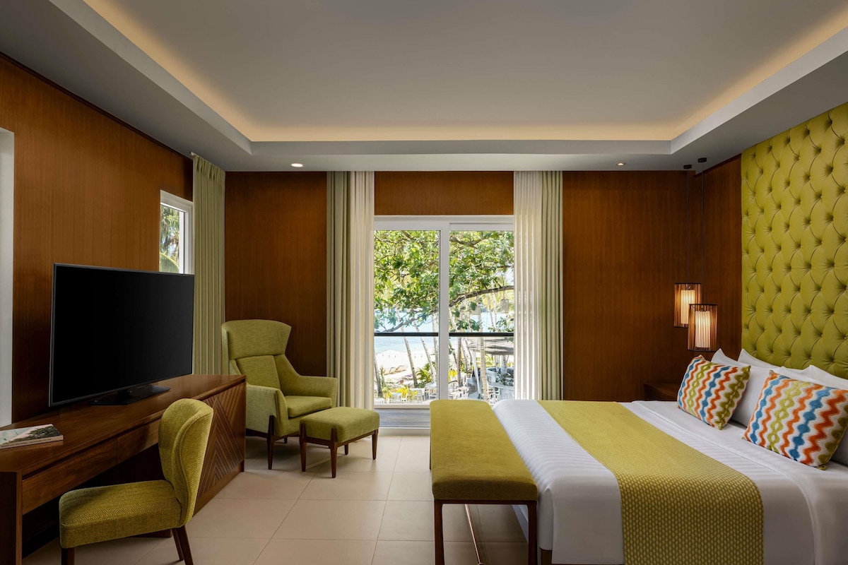 A hotel room with a bed, a desk with a TV, a chair, a bench, and a view of the outdoors.