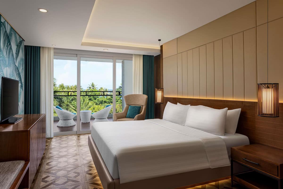 Habitación de hotel con una cama grande, un escritorio, una silla, un televisor y un balcón con vistas a zonas verdes.