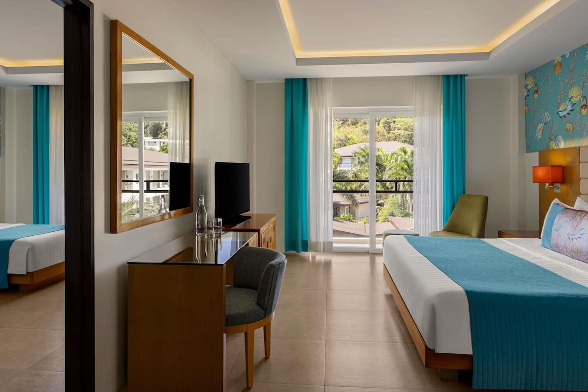 Habitación de hotel con cama, escritorio con televisor, una silla, un espejo grande y vistas al exterior.