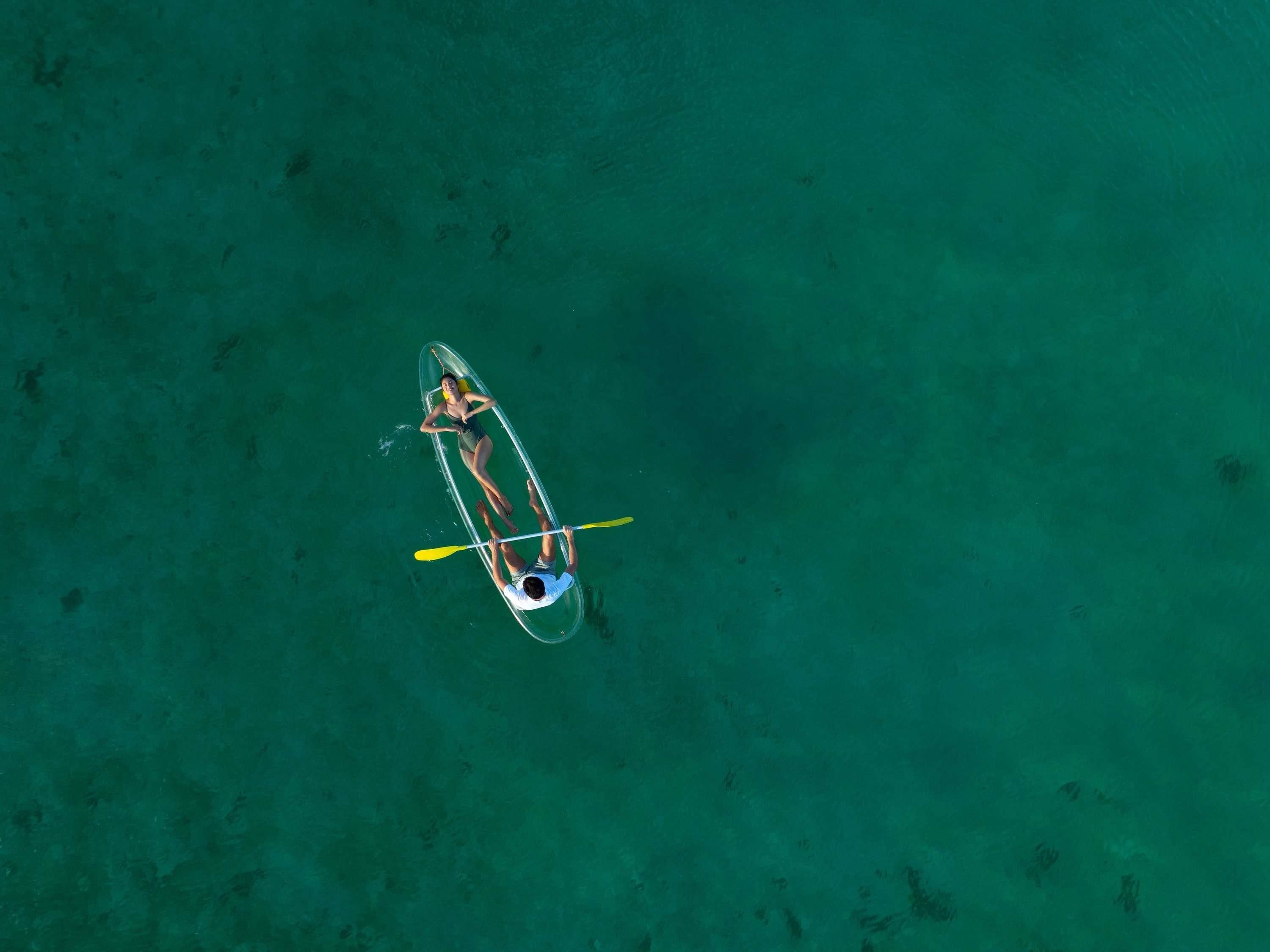 An aerial view of a clear turquoise body of water with a single transparent canoe containing two people.