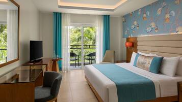 Habitación de hotel con cama, escritorio con televisor, una silla, balcón con sillas y vistas a zonas verdes.