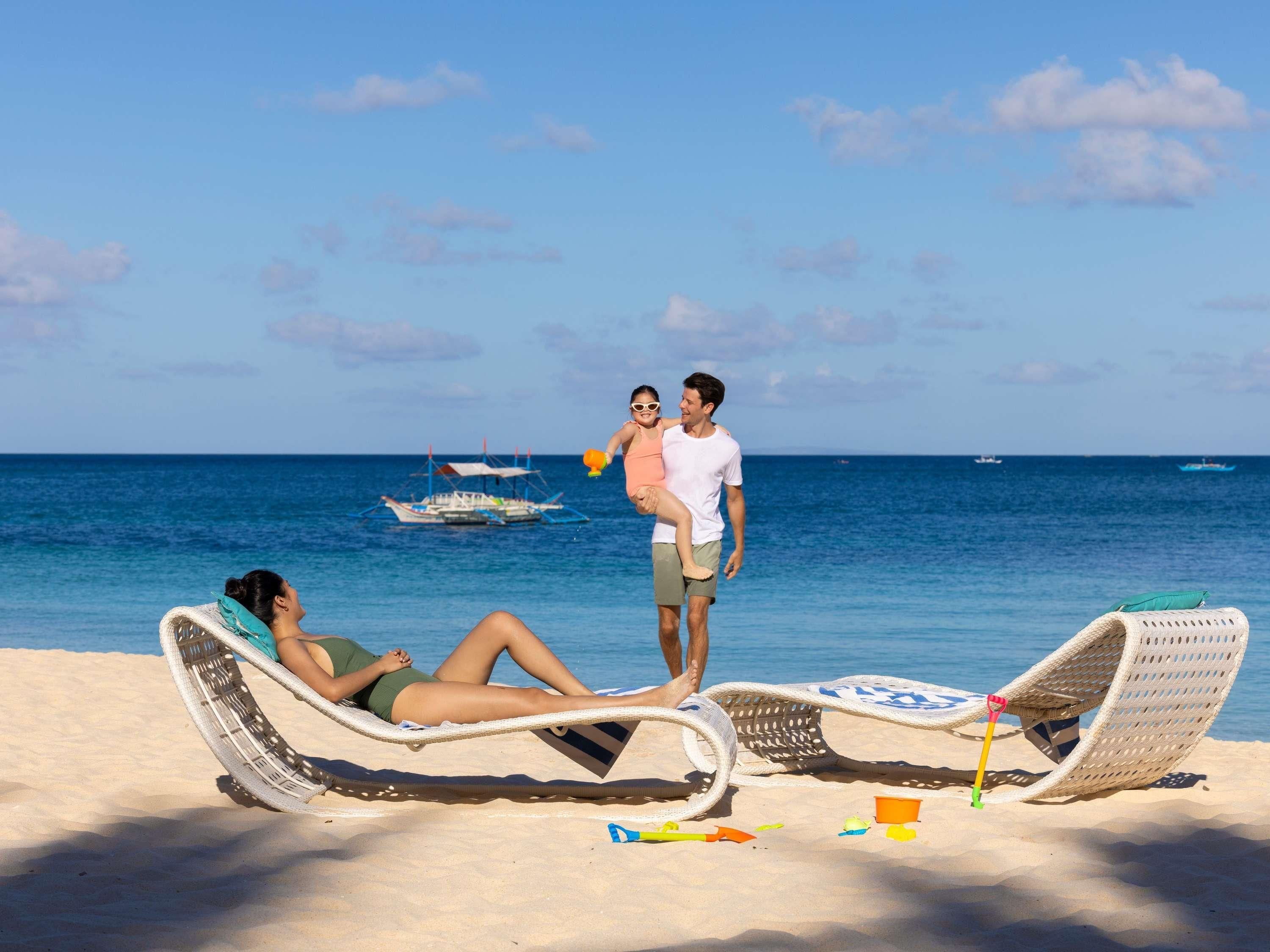 Escena de playa con dos sillas de playa, una persona acostada en una de ellas y otra de pie en la otra. Se ven barcos al fondo.
