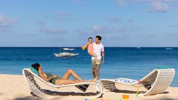 Escena de playa con dos sillas de playa, una persona acostada en una de ellas y otra de pie en la otra. Se ven barcos al fondo.
