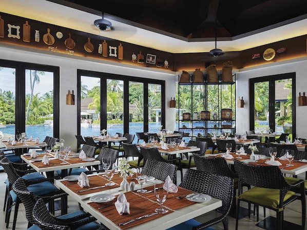 Un restaurant avec des tables dressées pour le dîner, de grandes fenêtres donnant sur une piscine et des palmiers, et un plafond orné de luminaires suspendus.