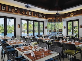 Un restaurante con mesas dispuestas para comer, ventanas amplias con vista a una piscina y palmeras, y un techo con luces colgantes.