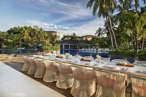 Ein Essbereich mit einem langen Tisch, gedeckt für eine Mahlzeit, umgeben von Korbstühlen, mit einem Pool und Palmen im Hintergrund.