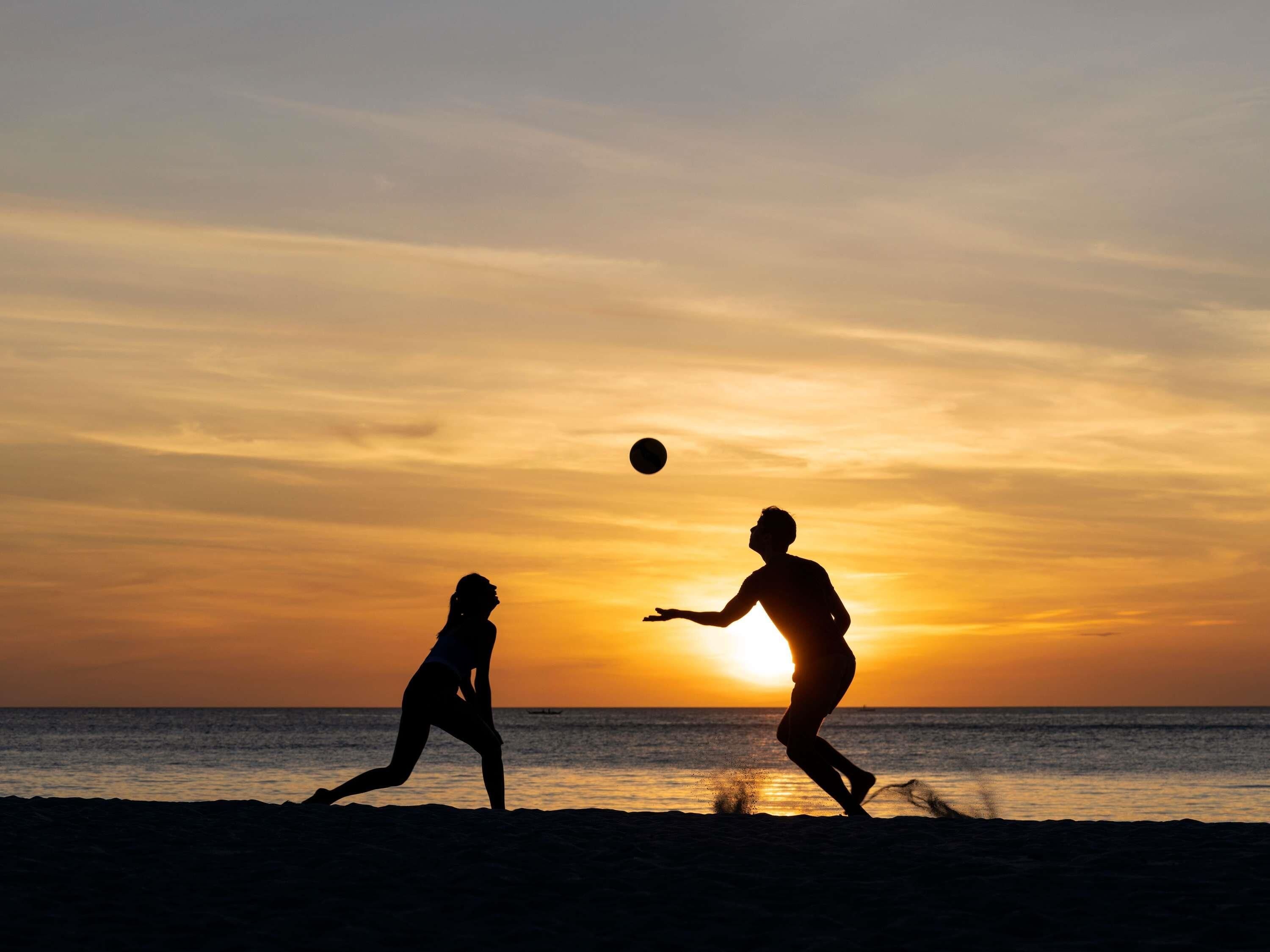 Silhouetten von zwei Personen, die am Strand bei einem Sonnenuntergang mit einem Ball spielen.