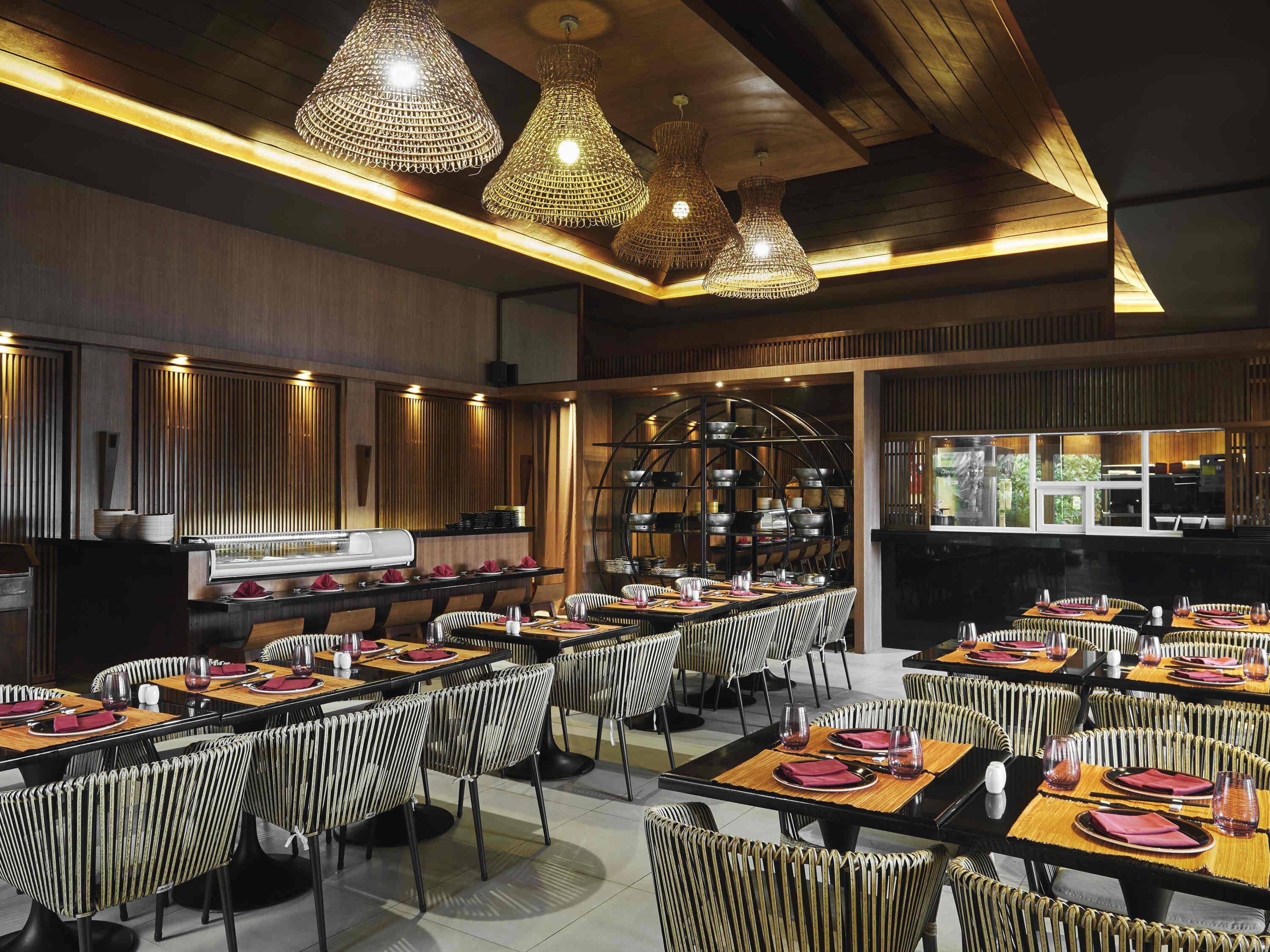 A restaurant with multiple tables, wicker chairs, and hanging light fixtures.