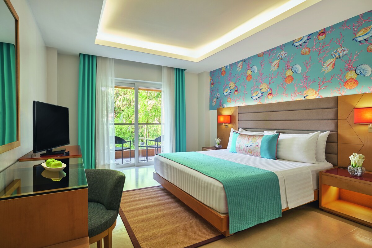 Een hotelkamer met een bed, een televisie, een bureau en een stoel. Er is een raam met gordijnen en uitzicht op groen buiten.