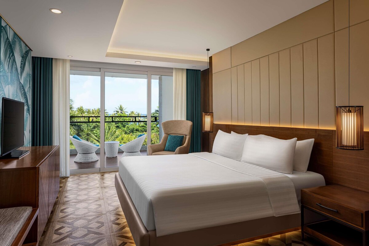 Une chambre d’hôtel avec un grand lit, un bureau, une chaise, une télévision et un balcon donnant sur un paysage verdoyant.