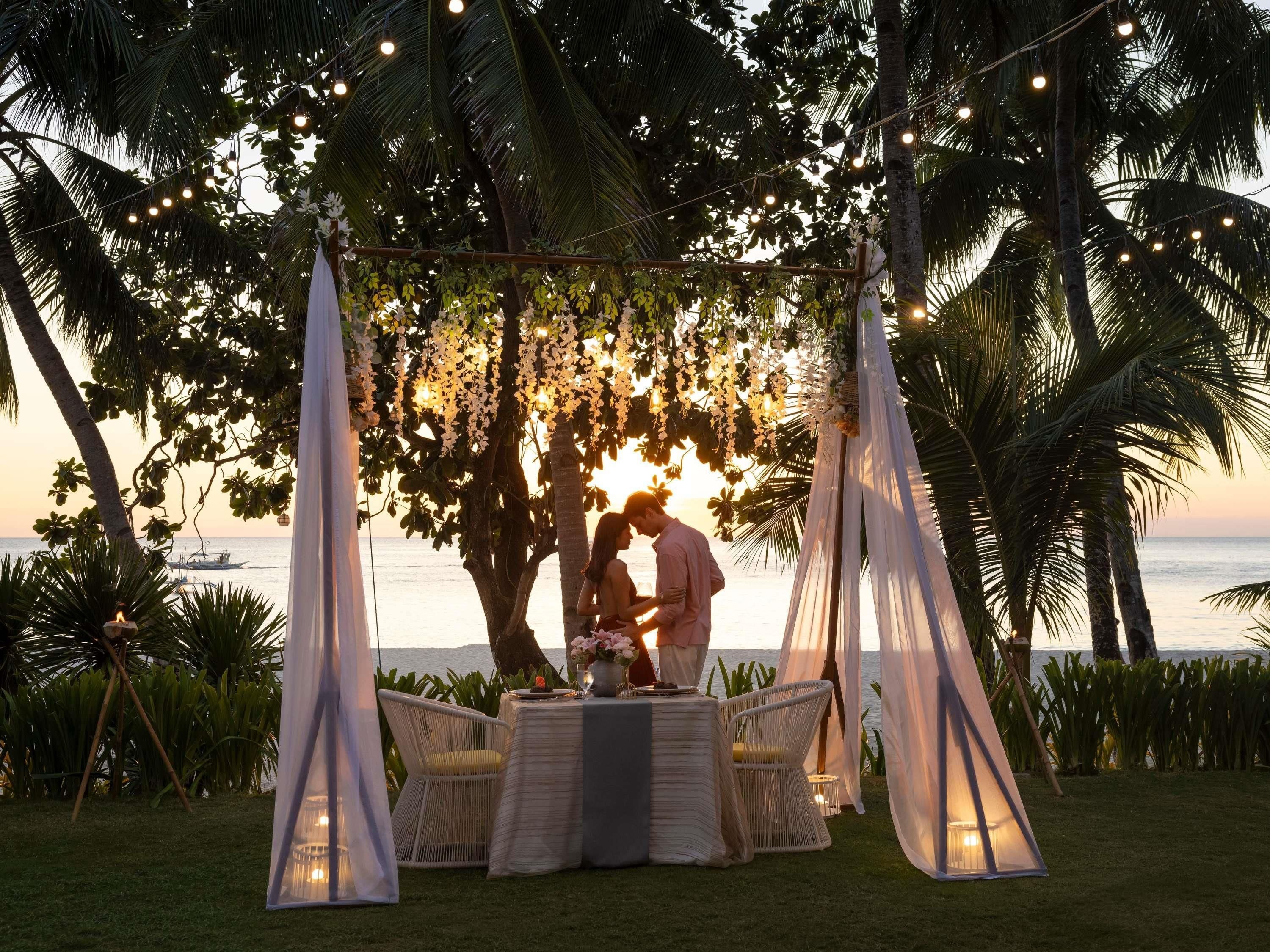 Ambiente tropical ao ar livre com toldo, luzes de corda e uma mesa com cadeiras.