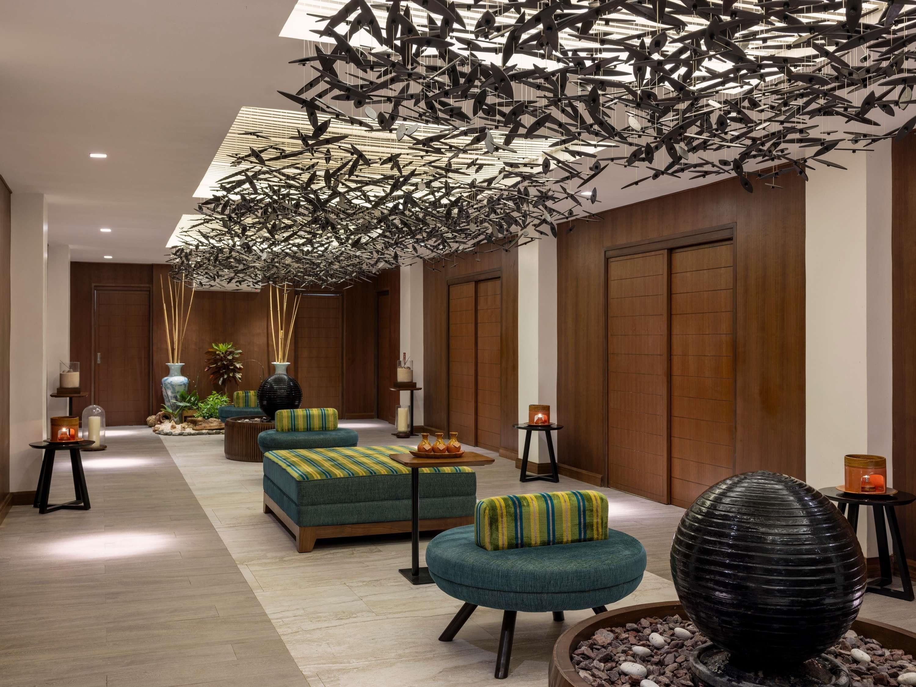 A modern lobby with a high ceiling, wooden paneling, and a unique ceiling installation of hanging elements. There are seating areas with cushions and a decorative water feature.