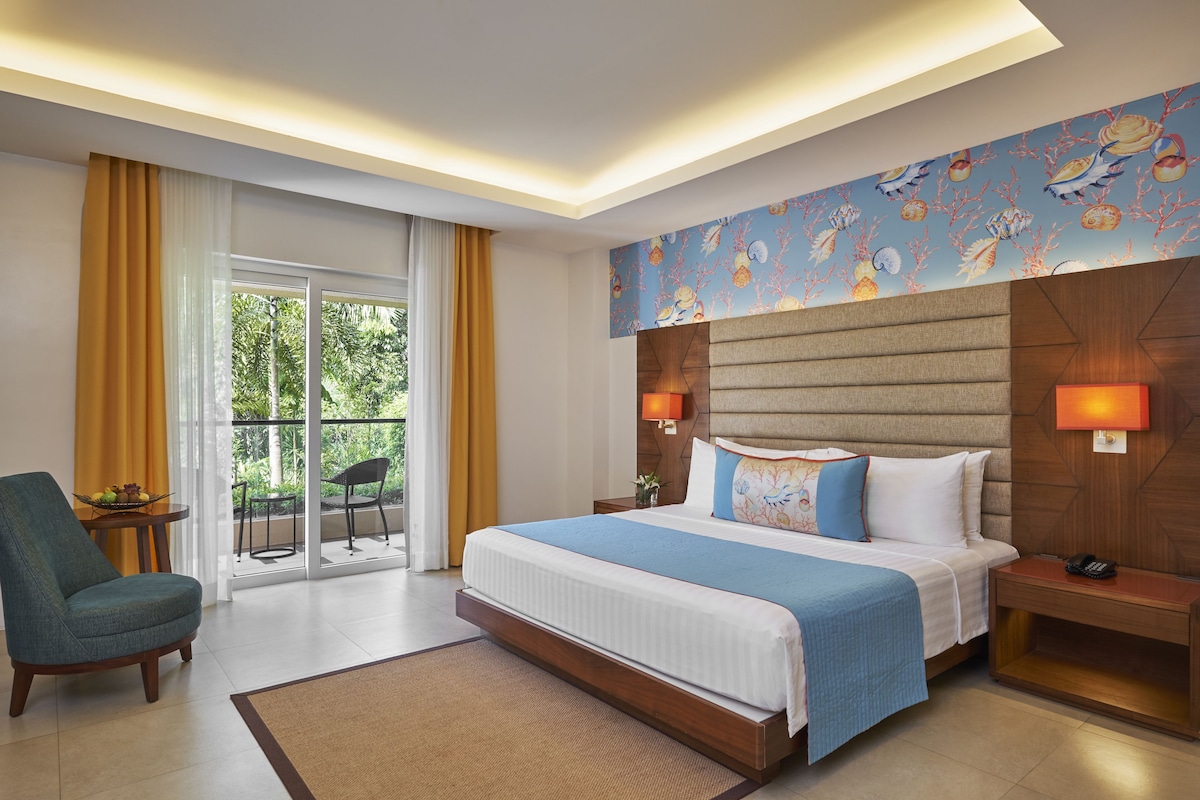 Een moderne hotelkamer met een groot bed, een zithoek met een stoel, een kleine tafel met fruit en een balkon met gele gordijnen.