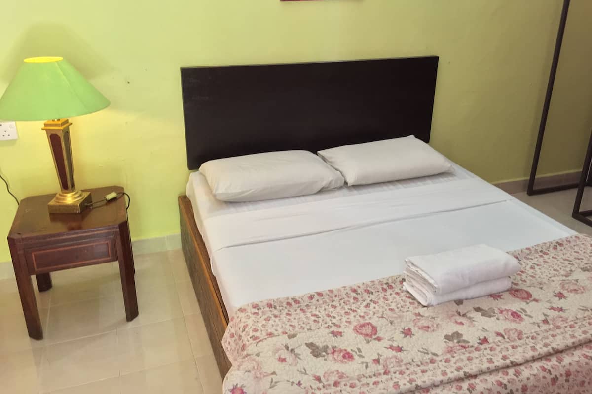 A neatly made bed with white linens, a floral-patterned blanket, and a folded towel on the bed. There is a small wooden nightstand with a green lamp beside the bed.