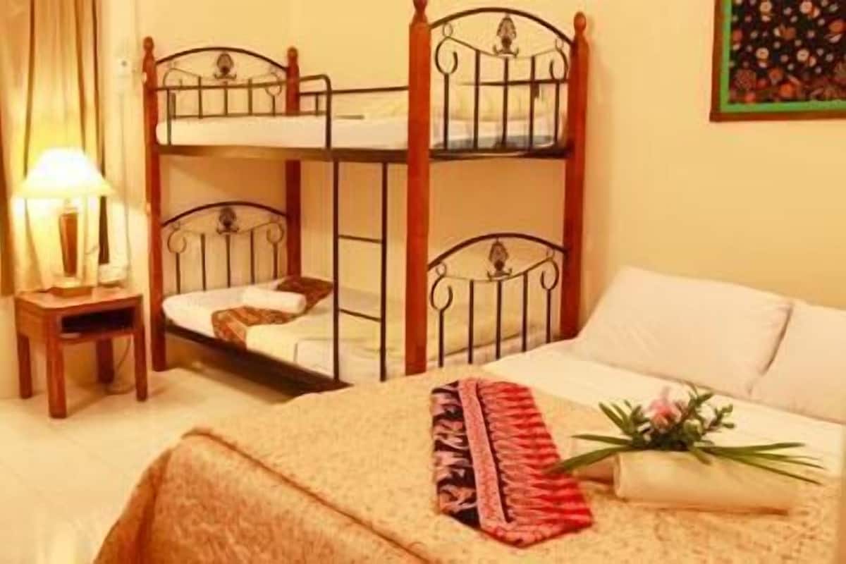 A hotel room with a bunk bed, a bedside table with a lamp, a bed with a floral arrangement, and a painting on the wall.