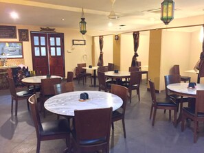 A restaurant with round tables and chairs, a kitchen in the background, and hanging lanterns.